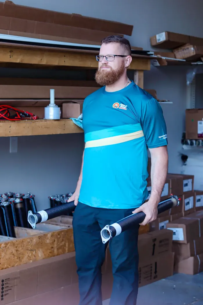 Good Day Garage Doors technician holding two garage door torsion springs inside a parts warehouse with shelves of inventory.