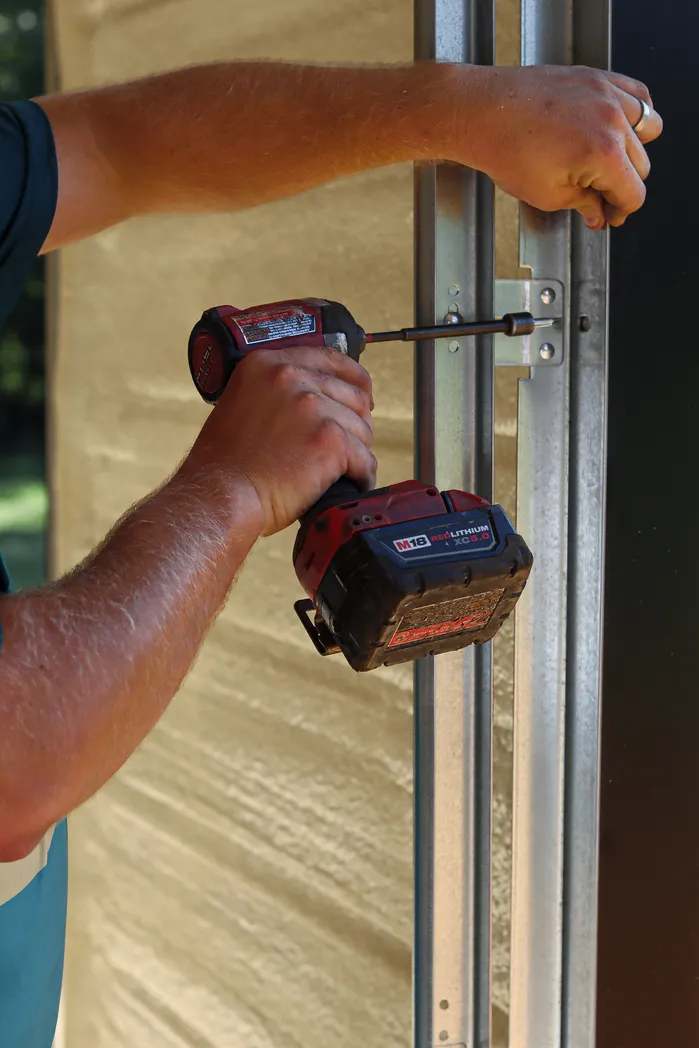 Close-up of a Milwaukee M18 cordless impact driver tightening hardware on a vertical galvanized garage door track.